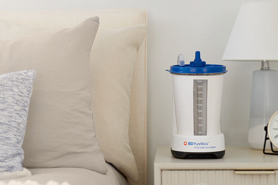 The PureWick™ Urine Collection System sits on a nightstand beside a bed and lamp